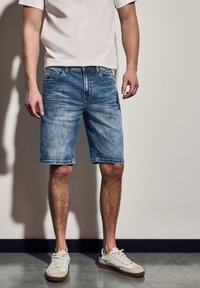 Jeansshorts i ljusblå tvätt, med klassisk femfickdesign, rå fåll och subtilt blekt textur. Bärs med vita sneakers.