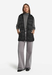 Czarna, pikowana kamizelka z kapturem, przednimi kieszeniami i zamkiem błyskawicznym. W połączeniu z szarym golfem i jasnoszarymi spódnicami dzwonami. Gładka tekstura.