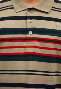Μπεζ πόλο πουκάμισο με οριζόντιες κόκκινες, πράσινες και μαύρες ρίγες, τρία κουμπιά και κεντημένα αρχικά γράμματα "MN" στο στήθος.