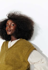 Gilet en polaire texturé vert olive avec un décolleté en V, porté sur une chemise à rayures légère à manches longues ; le mannequin arbore des cheveux naturels et volumineux.