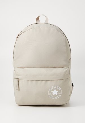 Zaino beige realizzato in tessuto resistente, con una forma arrotondata, una tasca anteriore con zip e una toppa con logo bianco sul fondo.