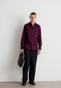 Burgunderfarget skjorte med knapper, sorte bukser, svarte sko, holder en svart dokumentmappe. Glatt stoff, strukturert design, minimalistisk stil.