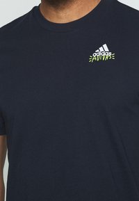 Tmavě modré bavlněné tričko s krátkými rukávy. Obsahuje bílé logo Adidas a neonově zelené akcenty poblíž loga, s hladkou texturou.