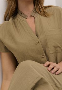 Blusa color khaki con colletto arricciato, maniche corte e chiusura con un solo bottone. Presenta una tasca frontale e un tessuto morbido e testurizzato.