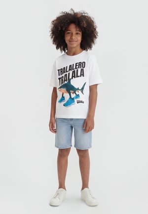 Παιδί με σγουρά μαλλιά φοράει λευκό μπλουζάκι με ένα καρχαρία με μπλε αθλητικά παπούτσια και κείμενο, ανοιχτόχρωμα τζιν σορτς και λευκά αθλητικά παπούτσια.