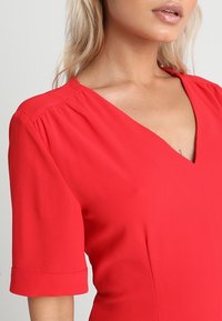 Röd klänning med V-urringning och korta upprullade ärmar. Tillverkad av textil med struktur, med mjuka draperingar och en figursydd silhuett.