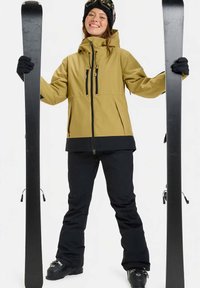 Lächelnde Person in gelber Jacke und schwarzen Hosen hält zwei schwarze Skier aufrecht, trägt Handschuhe, Skischuhe, Skibrille und eine schwarze Mütze.