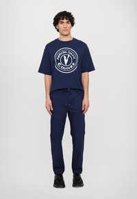 Zils oversized T-krekls ar balto logo druku, kombinēts ar zilajiem jogger biksēm ar elastīgu jostu un sānu kabatām. Melni sporta apavi.