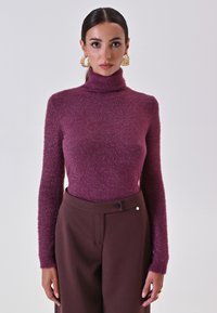 Donna con i capelli scuri, indossante un maglione viola a collo alto e maniche lunghe, insieme a pantaloni marroni a vita alta, con grandi orecchini a cerchio dorati.