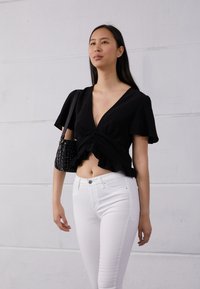 Top preto de manga curta e cropped com decote em V profundo e bainha com babado, combinado com jeans skinny brancos e uma mala preta texturizada.