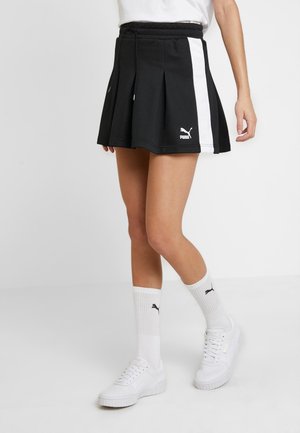 Jupe plissée noire en mélange de coton, avec des rayures blanches sur les côtés et une taille élastique à cordon. Présentée avec des baskets blanches et des chaussettes.