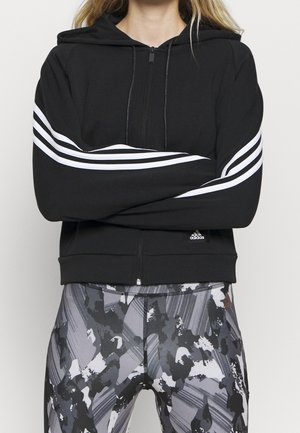 Person iført en sort hættetrøje med lynlås og hvide striber på ærmerne samt sorte, hvide og grå leggings med abstrakt mønster, med armene krydset.