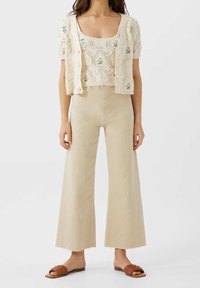 Beige knitted two-piece outfit featuring a short-sleeve cropped cardigan with floral details and wide-leg high-waisted pants. Flat sandals.