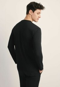 Czarna bluzka z długim rękawem wykonana z gładkiego materiału, z okrągłym dekoltem i dopasowanym krojem, z delikatnym strechowym wykończeniem. Widok z tyłu.