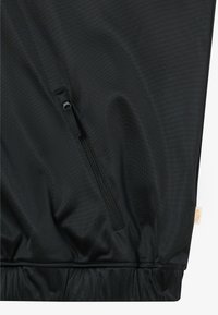 Černá texturovaná větruodolná bunda s kapsou na zip na přední straně a elastickým lemem. Obsahuje malou značkovou etiketu v dolním pravém rohu.
