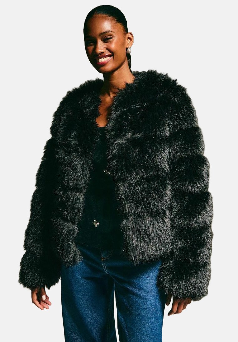 Mujer sonriendo con una chaqueta de piel sintética negra sobre una camiseta negra y jeans azules, de pie contra un fondo claro y liso.