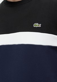 Gros plan d'une chemise noire, blanche et bleu marine avec un petit logo crocodile vert brodé sur la partie noire près de la poitrine.