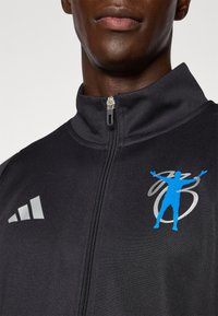 Črna športna zip-up jakna z sivimi akcenti, ki ima modro grafično logo osebe z dvignjenimi rokami. Gladka tekstura, visok ovratnik.