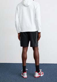 Personne debout sur un tapis bleu, portant un sweat à capuche blanc, un short noir à bandes blanches, des chaussettes noires et des baskets argentées et rouges, vue de dos.