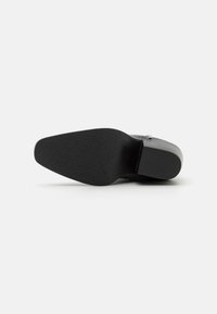 Zapato de tacón negro con suela de goma texturizada, que presenta un material superior liso y un diseño de punta cuadrada, acentuado por una hebilla lateral.
