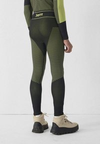 Zwart met neon groene gestreepte legging met een hoge tailleband waarop de merknaam "OTT" staat. Gladde textuur, aansluitend ontwerp; gecombineerd met beige wandelschoenen.