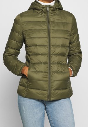 Personne portant une veste matelassée vert olive avec capuche, fermeture éclair frontale et mains dans les poches, associée à un jean bleu clair.