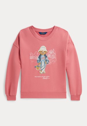 Sweat-shirt rose avec un ours portant un chapeau, une veste en jean, un pantalon rayé, tenant un panier, avec des croquis de maison et de jardin en arrière-plan.