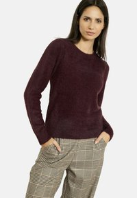 Maglione borgogna, peloso, con scollatura rotonda e maniche lunghe, abbinato a pantaloni a quadri beige e marroni.