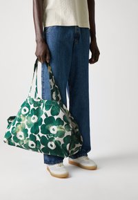 Tasche mit grünem Blumenmuster aus Baumwollstoff, ausgestattet mit zwei stabilen Griffen und einem geräumigen Design, gehalten von einer Person.