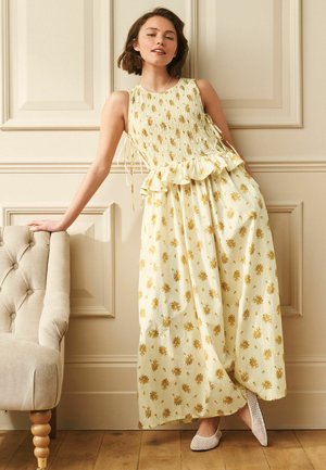 Junge Frau in einem ärmellosen gelben Blumenkleid und weißen Netzschuhen lehnt an einem beigefarbenen, geknöpften Sessel vor vertäfelten Wänden.