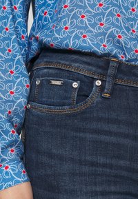 Mörk denimjeans med orange sömmar och silverdetaljer, kombinerat med en blå blommig blus med lång ärm och röda accenter.