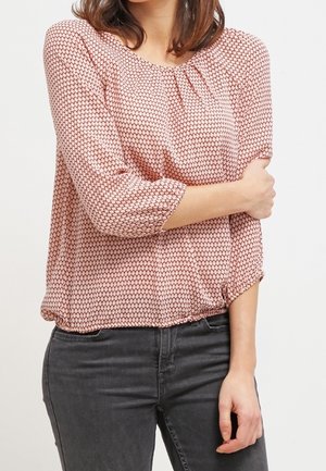 Femme portant un chemisier rose à motifs avec manches trois-quarts et un jean gris foncé sur un fond clair uni.