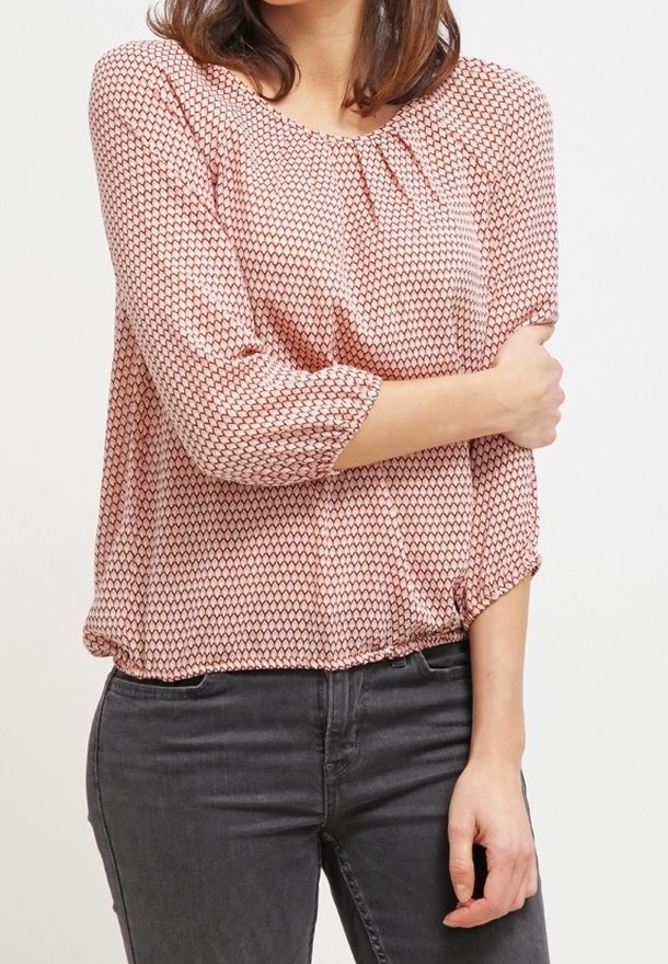 Femme portant un chemisier rose à motifs avec manches trois-quarts et un jean gris foncé sur un fond clair uni.