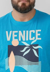 Turquoise T-shirt met het woord "VENETIË" in vetgedrukte witte letters, met grafische elementen zoals lijnen, een abstract figuur en plantvormen.