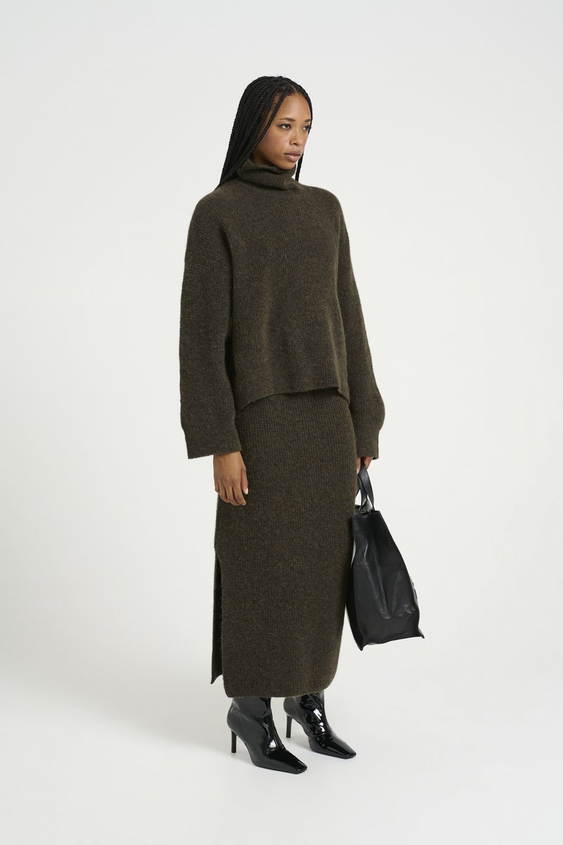 Maglione a collo alto in maglia verde oliva abbinato a una gonna aderente coordinata, con fessure laterali. Modella che tiene una borsa a tracolla in pelle nera.