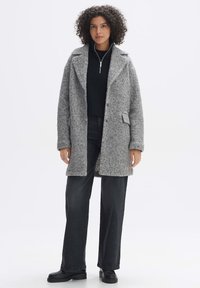 Frau mit lockigem Haar, die einen strukturierten grauen Mantel über einem schwarzen Reißverschluss-Sweater, dunklen weit geschnittenen Jeans und schwarzen Plateauschuhen auf weißem Hintergrund trägt.