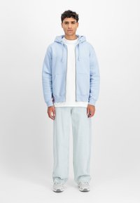 Világoskék cipzáras kapucnis felső oldalsó zsebekkel, fehér pólóval párosítva, világosszürke farmer széles lábakkal és szürke sportcipőkkel.