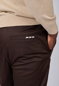 Homme portant un pull beige et un pantalon marron foncé avec trois logos de buffles blancs au-dessus de la poche arrière droite, la main dans la poche.
