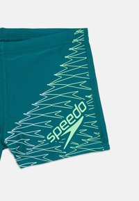 Tamsiai žali maudymosi šortai su neoninės žalios zigzagu ir "Speedo" logotipu ant kojos, pateikti prieš baltą foną.