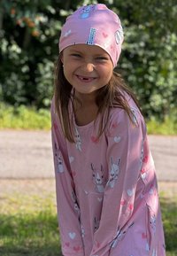 Vaaleanpunainen pitkähihainen mekko, jossa on pupu- ja sydänkuvioita, pehmeää kangasta. Sopiva vaaleanpunainen pipo, jossa on samankaltaiset kuviot. Hymyilevä lapsi ulkona.