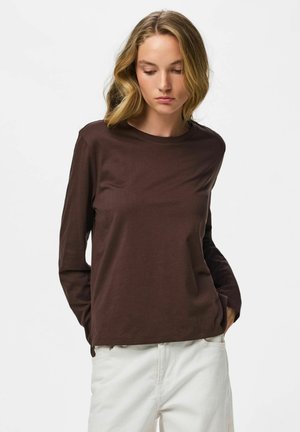 Jeune femme aux cheveux blonds ondulés portant une chemise marron à manches longues et un pantalon blanc, les mains dans les poches, regardant vers le bas avec une expression neutre.