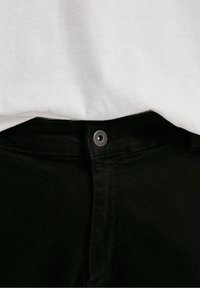 Gros plan d'un jean noir boutonné à la taille avec une partie d'une chemise blanche rentrée au-dessus de la ceinture.
