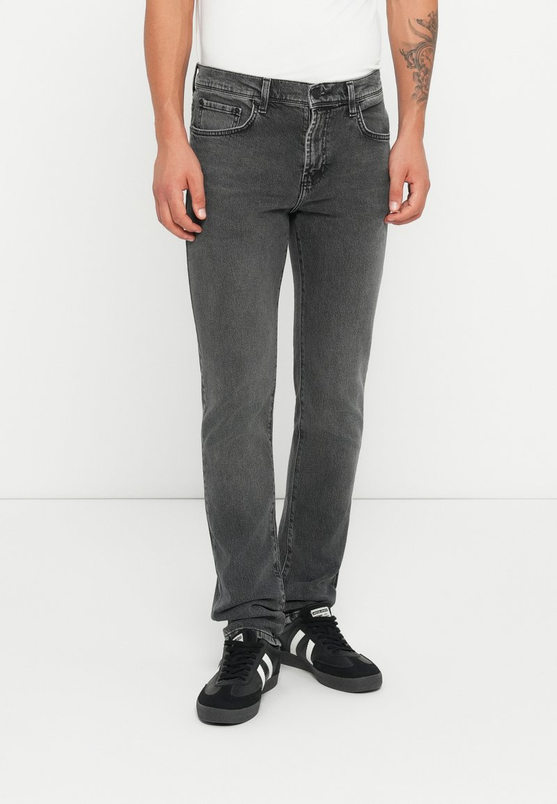 Άνδρας φορώντας εφαρμοστά γκρι τζιν και μαύρα αθλητικά παπούτσια με λευκές ρίγες, που στέκεται σε έναν απλό λευκό φόντο.