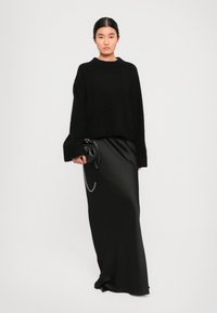 Maglione nero lavorato a maglia con scollatura a girocollo, abbinato a una lunga gonna nera in satin. La modella tiene una piccola borsa nera con una tracolla a catena.