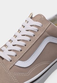 Μπεζ πάνινο αθλητικό παπούτσι με λευκή λωρίδα στο πλάι και χοντρά λευκά κορδόνια, με εμφανείς ραφές και μέρος της λευκής λαστιχένιας σόλας.