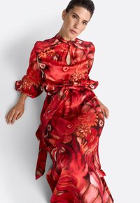 Röd blommig klänning i lätt tyg med nyckelhålsk neckline, voluminösa ärmar och midja med bälte för formgivning.