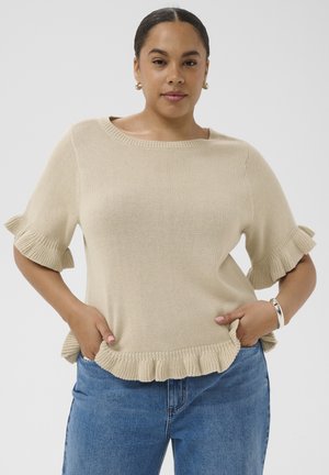 Femme aux cheveux foncés portant un pull en maille à manches courtes beige avec des détails à volants et un jean bleu, mains dans les poches, fond neutre.
