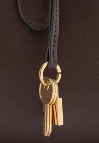 Hnedá kožená kabelka so zlatým krúžkom na kľúče, na ktorom sú tri kľúče a obdĺžnikový prívesok na kľúče. Stehy sú tmavo hnedé.