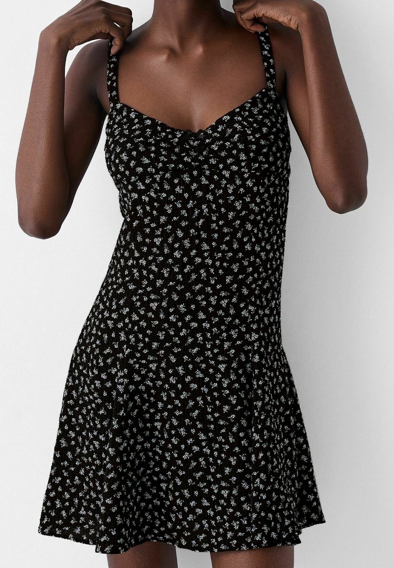 Svart klänning med små vita blommönster, figurnära bodice med justerbara axelband, utsvängd nederkant och textilstruktur.