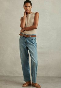Beige ribbede sleeveless top, blå højtaljede lige ben jeans, brun bælte med sølv spænde, og brune slidesandaler. Neutral baggrund.